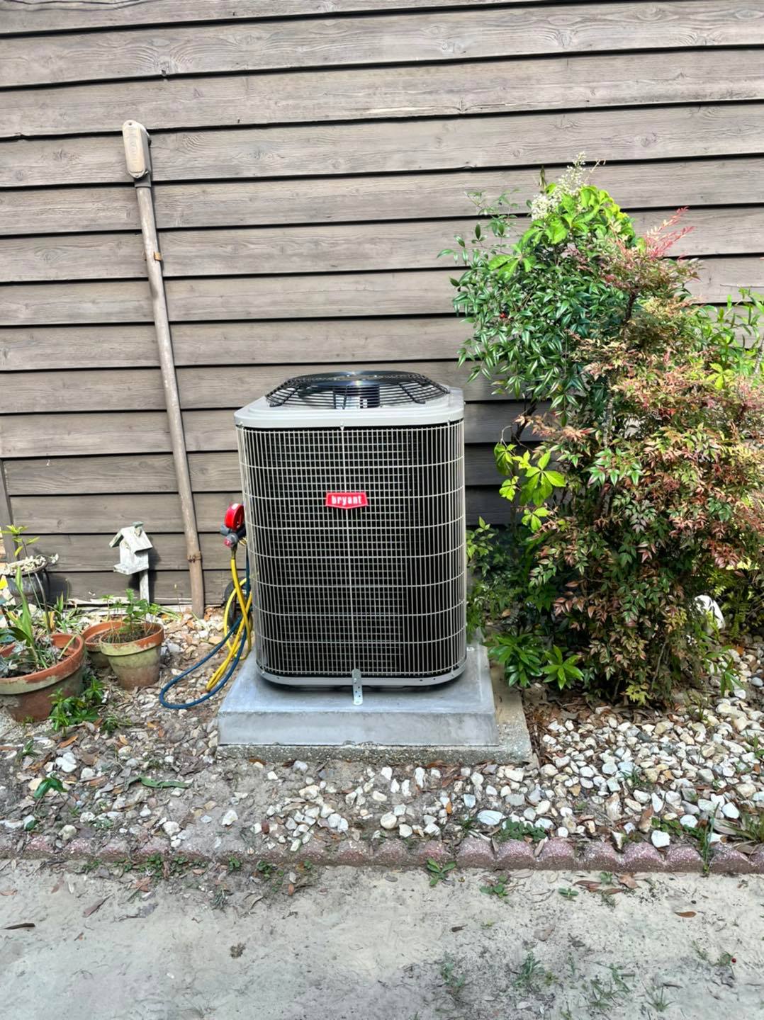 Outdoor Bryant air conditioning unit on concrete pad by house siding, surrounded by shrubs, potted plants and utility hoses