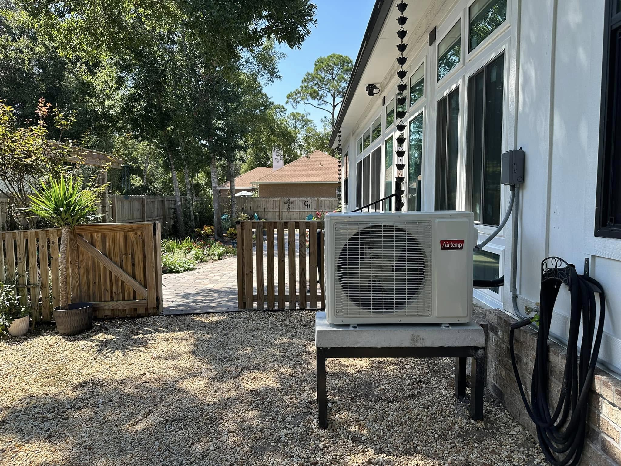 Outdoor Airtemp HVAC condenser by white house siding, gravel yard, wooden gate, rain chain and shaded backyard landscaping.