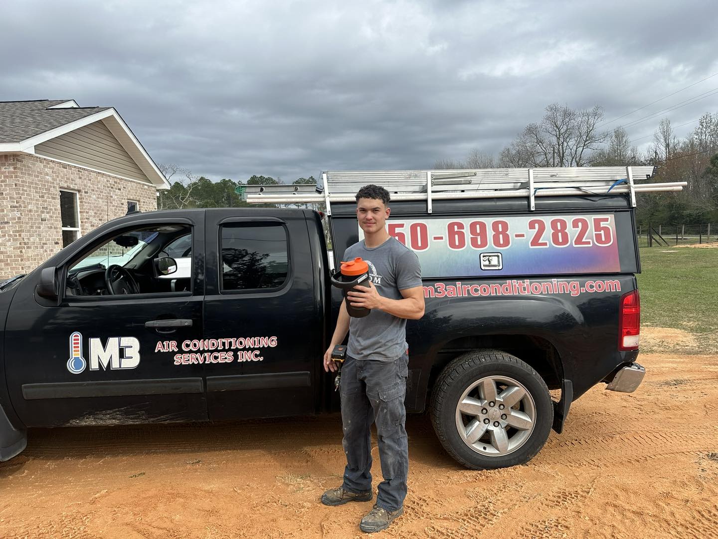 A man (HVAC technician) standing by a black M3 Air Conditioning service truck with ladder holding a cup and tools.
