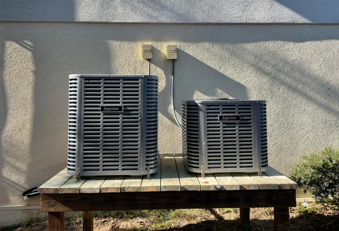 Two Ameristar outdoor air conditioning units on a wooden platform against a stucco wall.