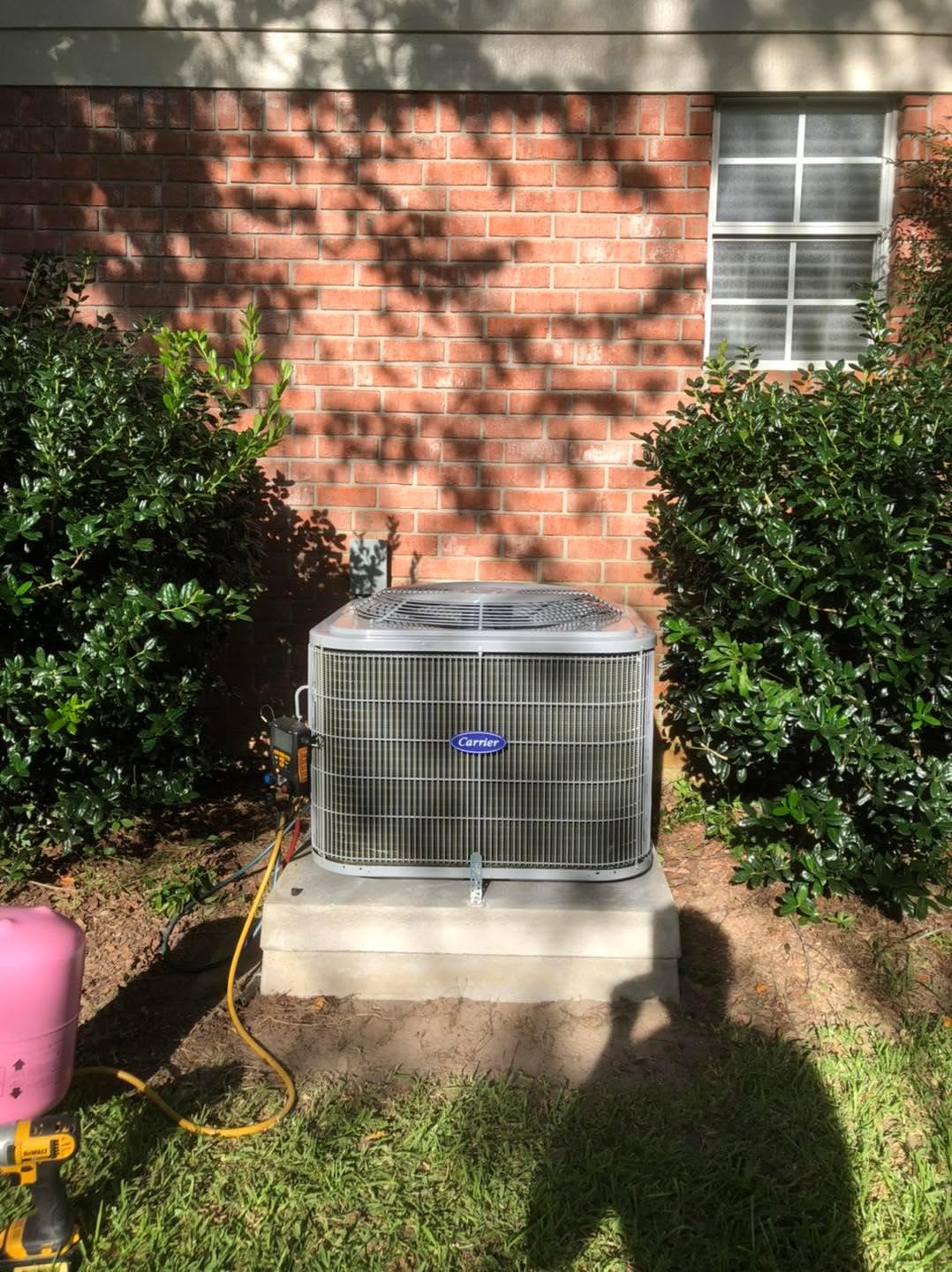 Carrier outdoor air conditioner on concrete pad by brick house, shrubs, service hoses and a person's shadow in foreground
