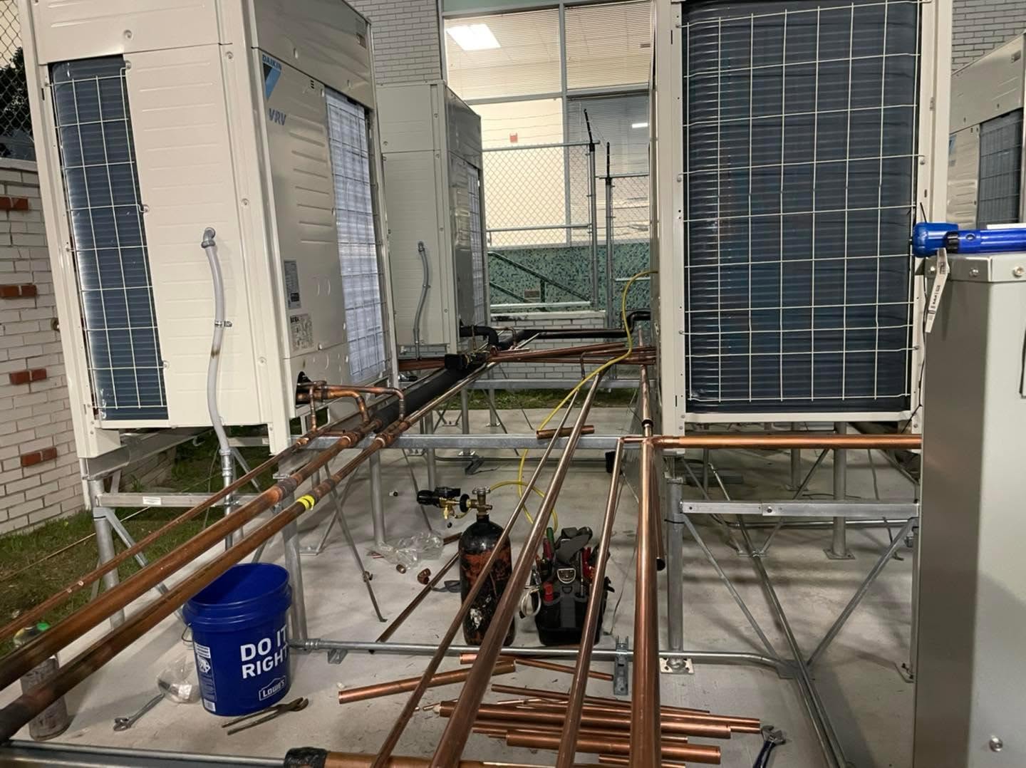 Copper refrigerant pipes connected to outdoor VRV air conditioning units on metal racks, tools and blue bucket nearby