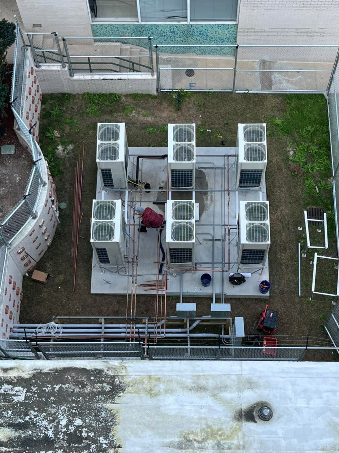 Aerial view of a person working among multiple rooftop HVAC units and copper piping in a fenced equipment yard.