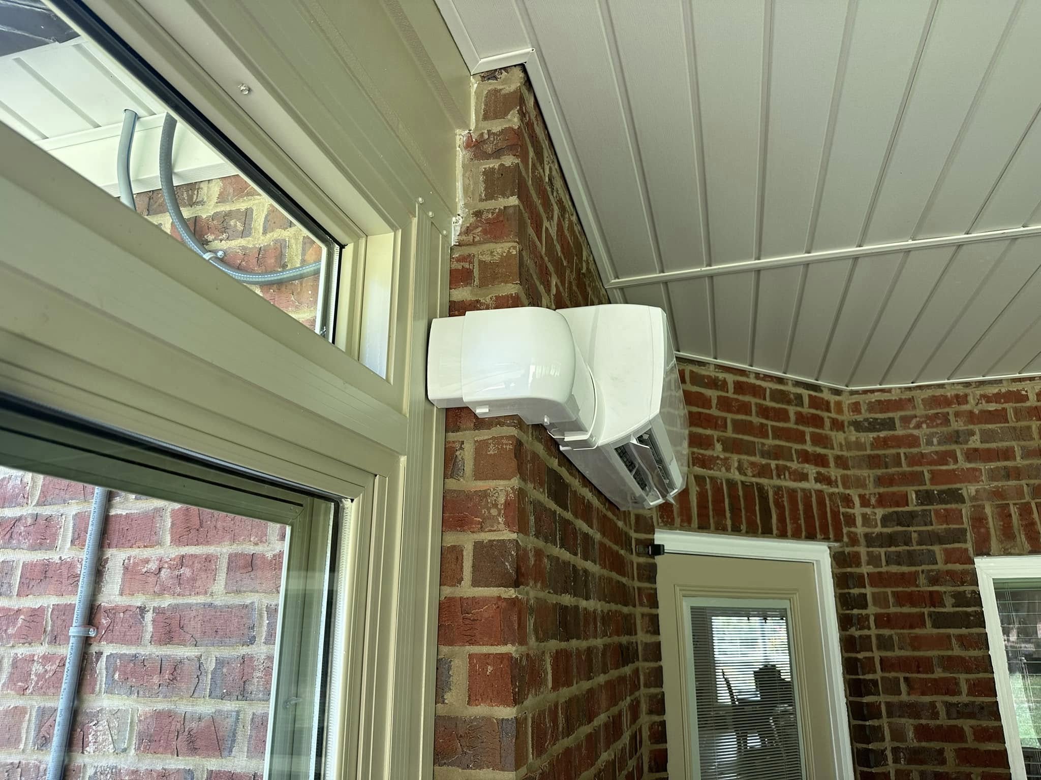 Ductless mini-split indoor unit mounted on brick interior corner beneath white PVC paneled ceiling near window