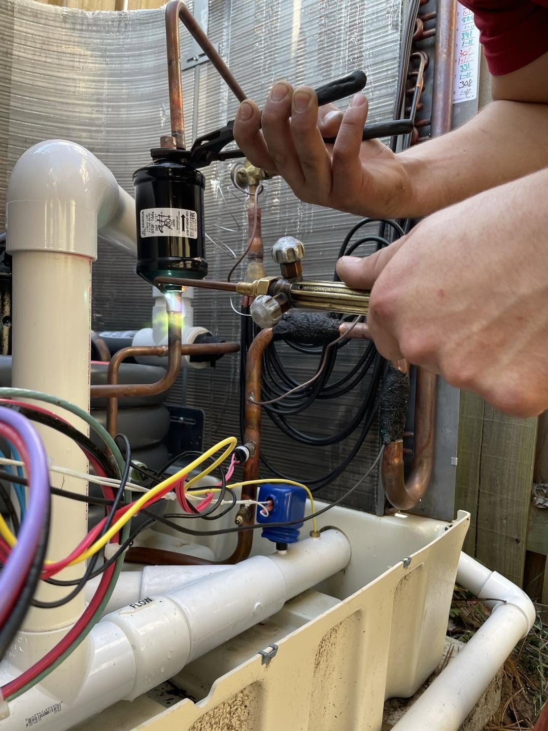 A person brazing copper refrigeration lines with torch and pliers inside an HVAC unit, wires and PVC drain visible