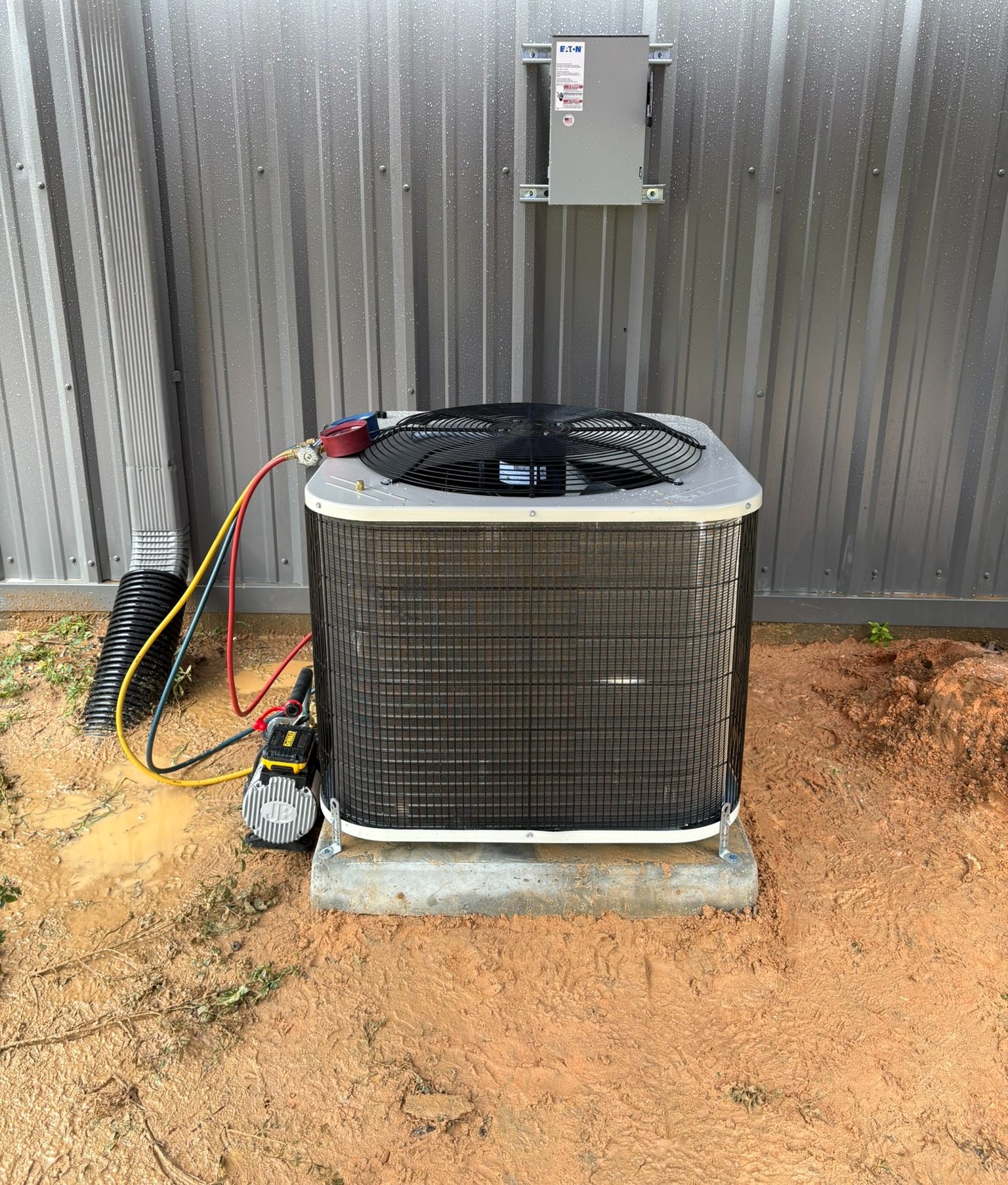Outdoor air conditioning condenser with service gauges and hoses beside metal building and electrical disconnect.