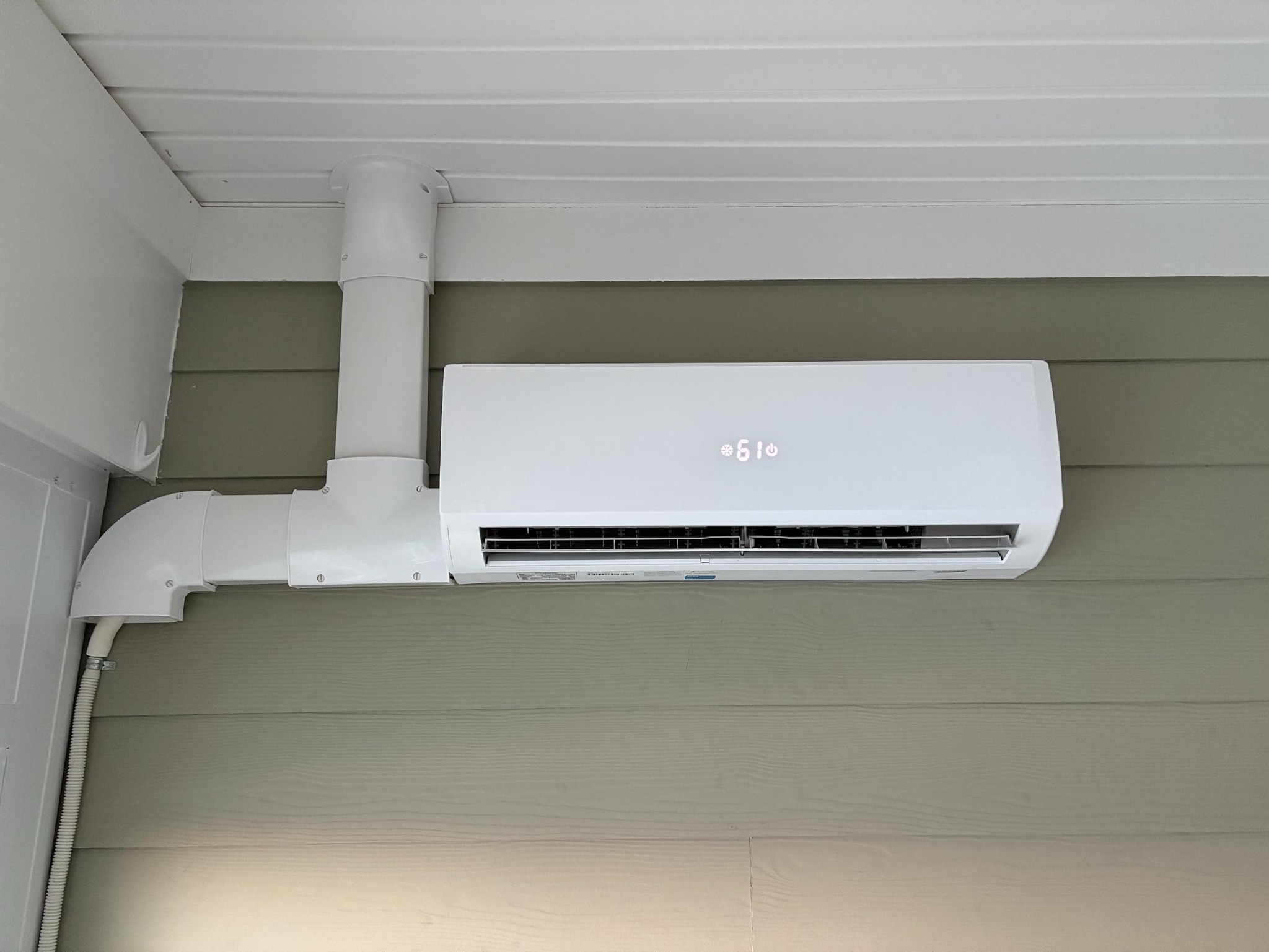 White wall-mounted ductless air conditioner with PVC ducting under a white ceiling on gray siding