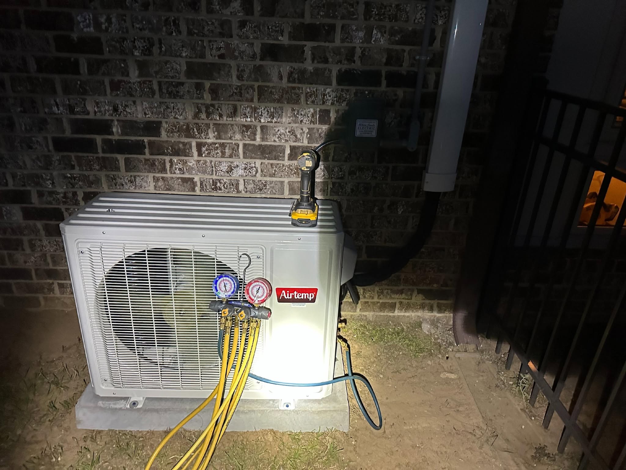 Outdoor Airtemp AC unit with HVAC manifold gauges and yellow service hoses against a brick wall at night.