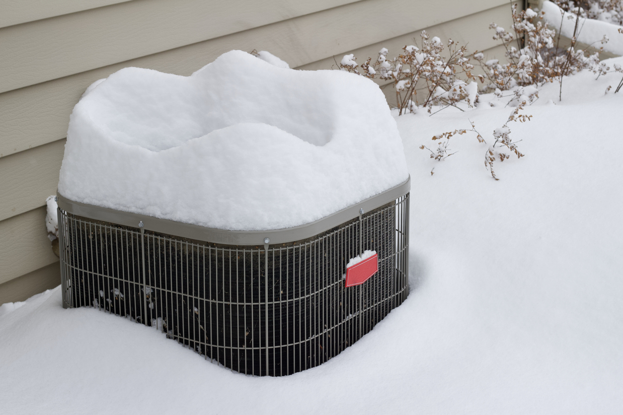 Outdoor air conditioning unit covered in deep snow beside house siding and snow-dusted plants