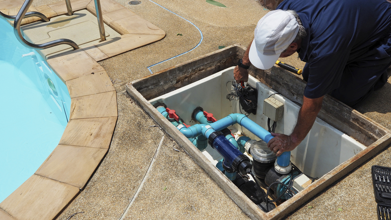 A man repairing blue PVC pool plumbing and filter components in an open equipment pit beside a swimming pool.