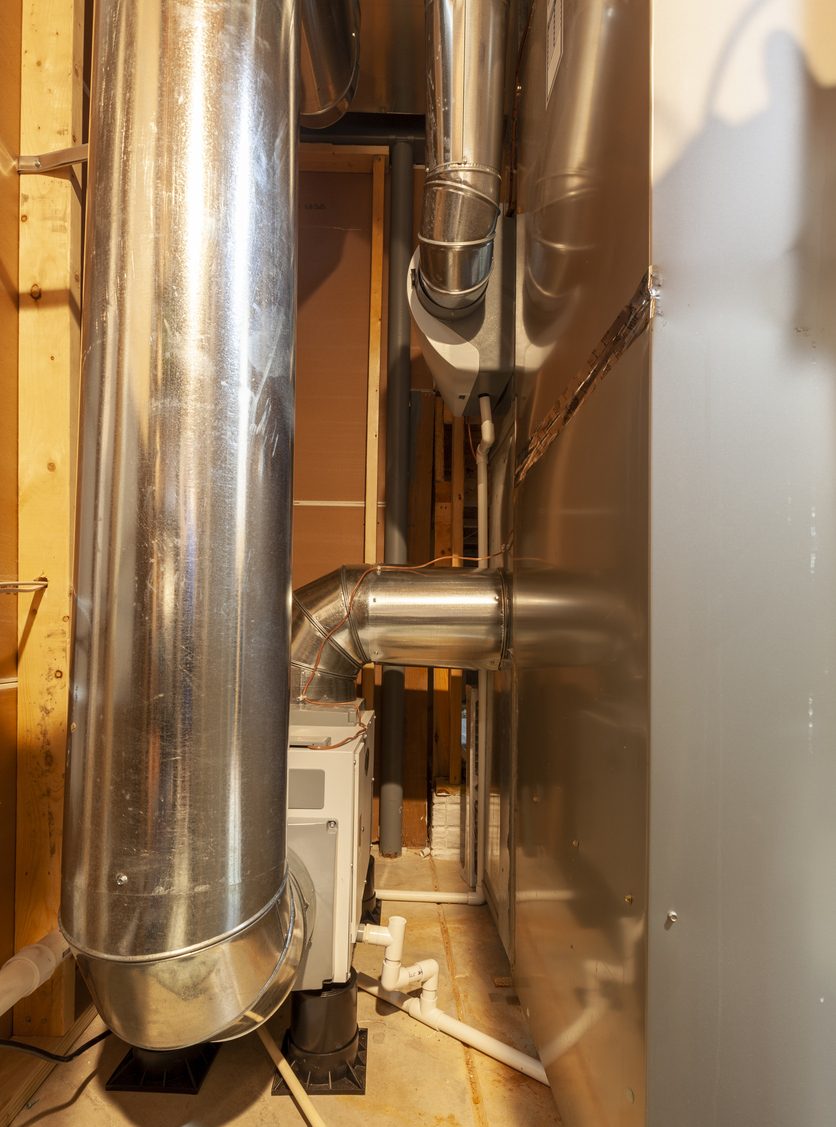 Utility room with metal HVAC ductwork, furnace unit, and PVC condensate drain pipes between wooden studs.