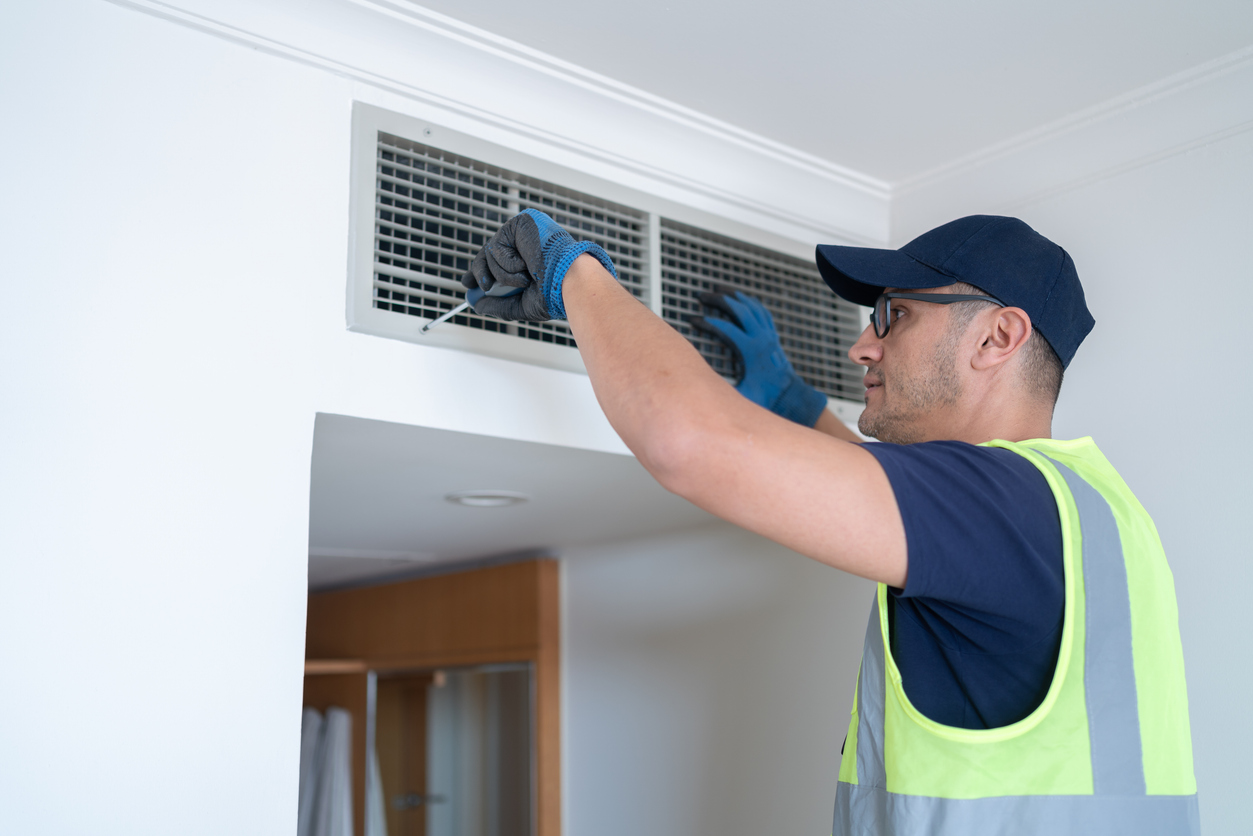 A man in a safety vest and cap fixing an indoor air vent grille with a screwdriver.